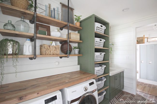 rustic laundry room with one shelving, shiplap, brick floor, green cabinets, laundry tower